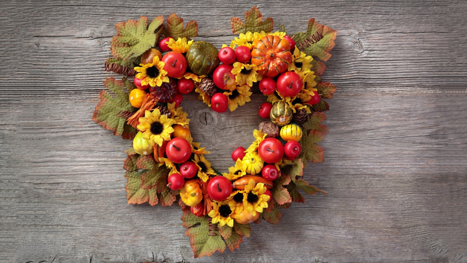 Heart-shaped fall wreath made of apples, mini pumpkins, sunflowers, and autumn leaves on a wooden background.