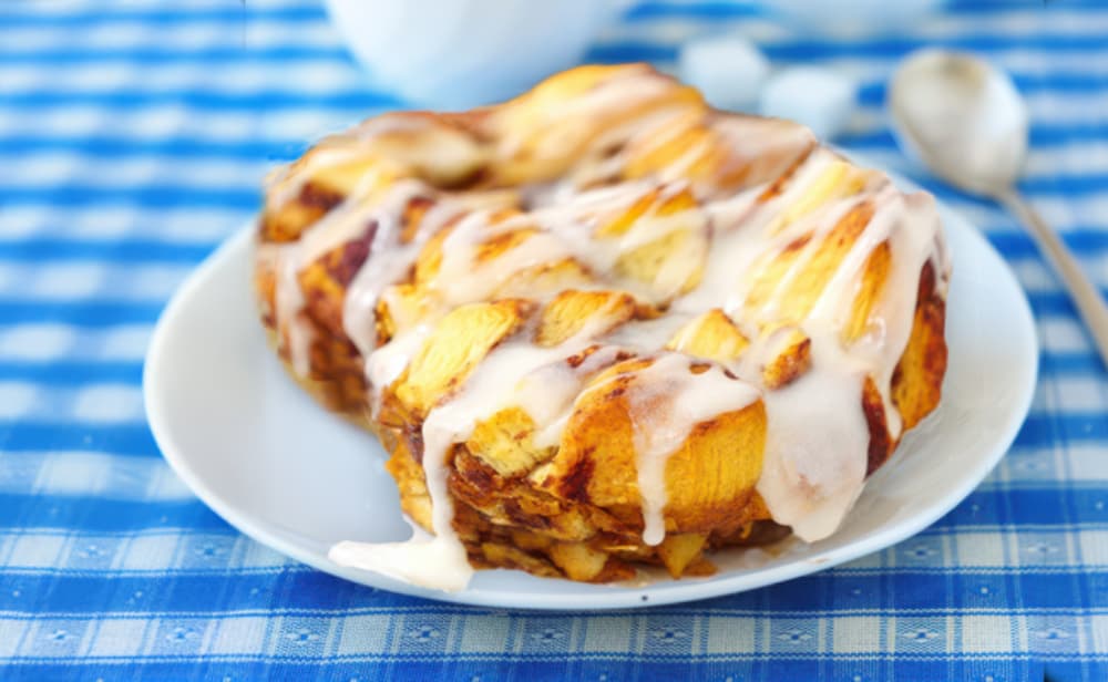 A warm serving of apple monkey bread topped with icing, sitting on a white plate over a blue checkered tablecloth.