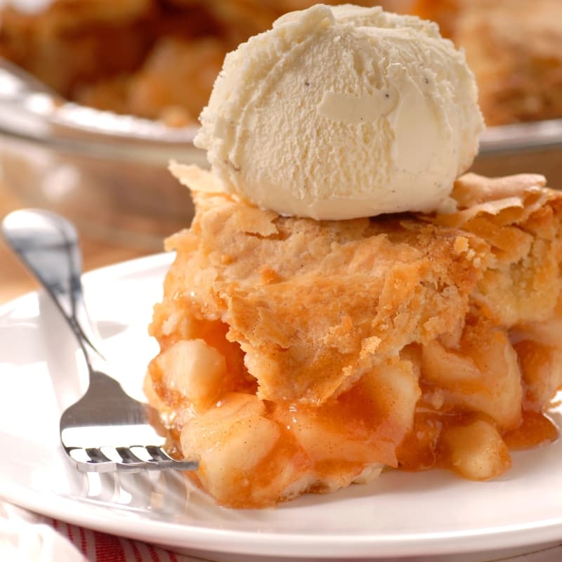 Close-up of a slice of apple pie topped with a scoop of vanilla ice cream on a white plate with a fork.
