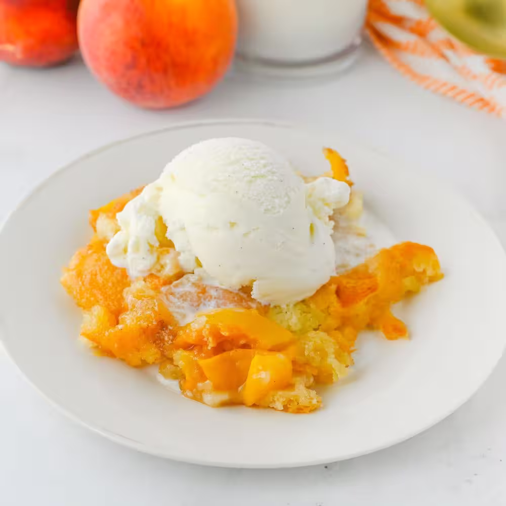 A close-up of a peach cobbler with vanilla ice cream, served on a white plate alongside whole peaches, with a glass of milk in the background.