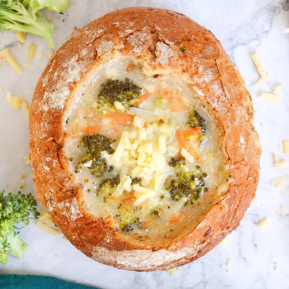 Overhead shot of broccoli cheddar soup served in a round bread bowl, topped with shredded cheese and surrounded by fresh broccoli.